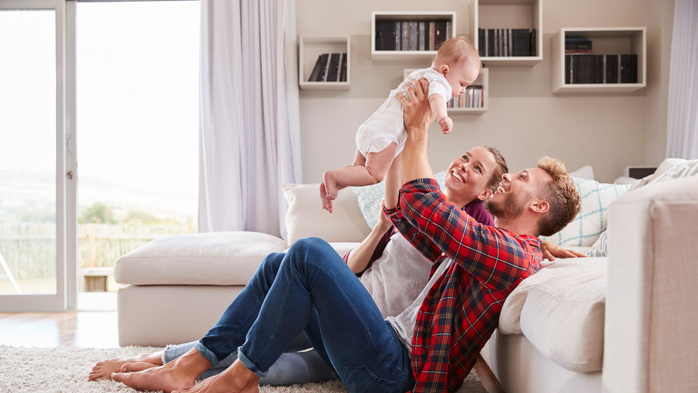 Junges Paar sitzt auf dem Teppich vor einem Sofa und hält fröhlich ein Baby in die Luft 